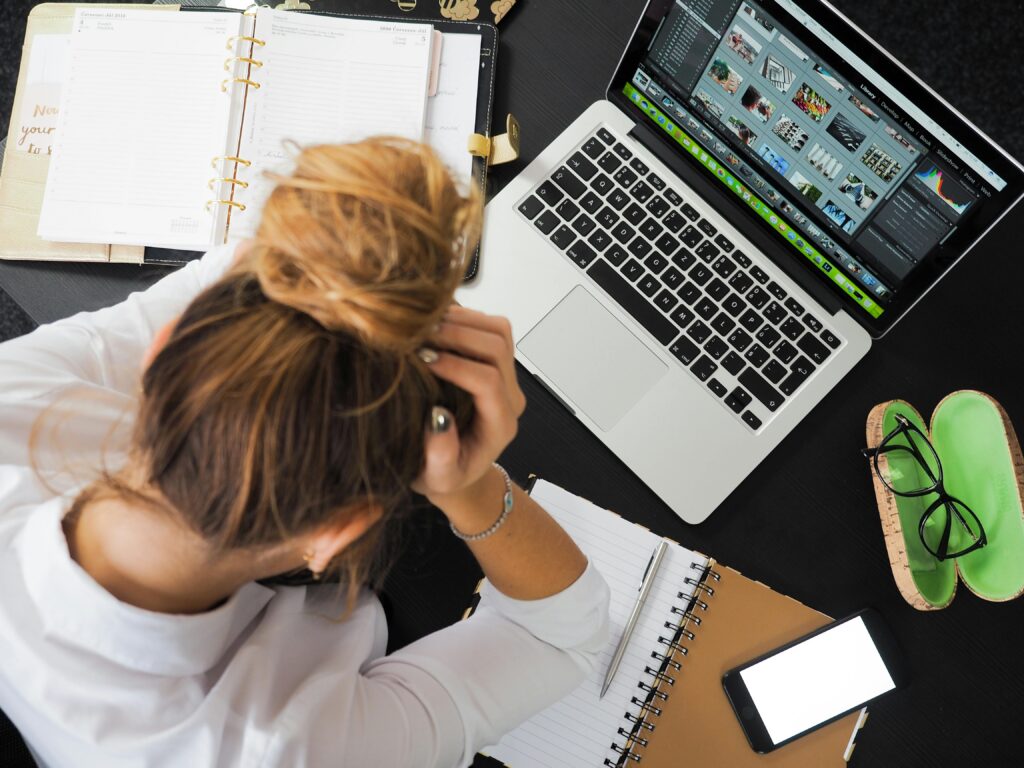 Stressed woman holding her head in front of a laptop, planner, and smartphone at a cluttered desk.