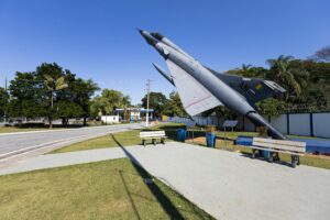 Military aircraft on display at a Brazilian air base, mounted upright beside a road near the entrance gate with benches and palm trees in view.
