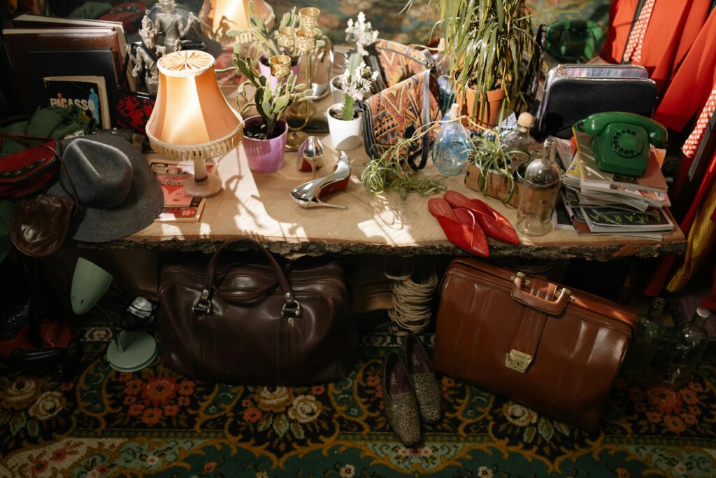 Vintage table setup with resale inventory including handbags, lamps, shoes, plants, magazines, and home décor on display in a cozy room.