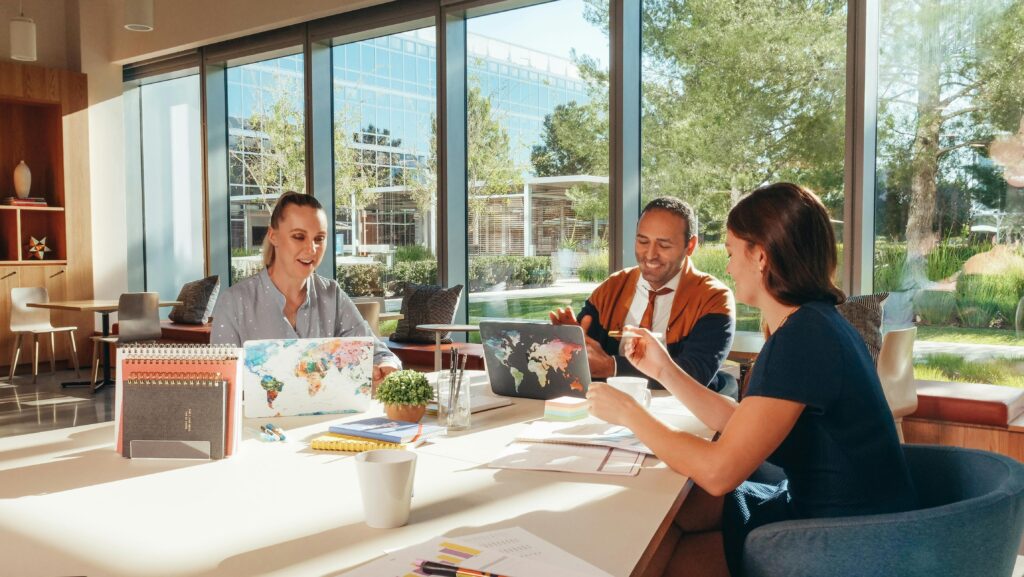 Three business professionals having a meeting at a sunlit office table with laptops, notebooks, and planners.