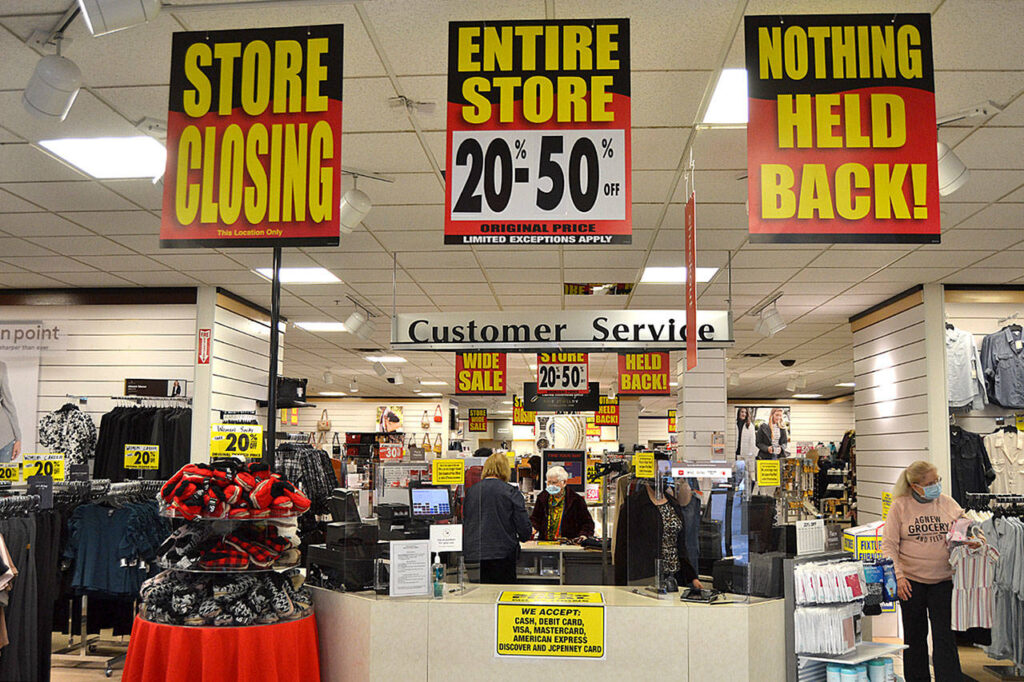 Inside a JCPenney store during liquidation with large yellow and red signs reading “Store Closing,” “Entire Store 20–50% Off,” and “Nothing Held Back,” as customers browse discounted merchandise.