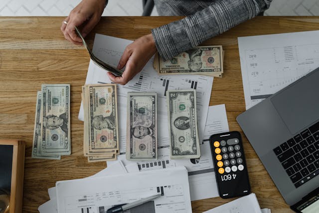 Person organizing U.S. dollar bills and financial documents at a desk with a calculator and laptop, planning expenses for life in a new country