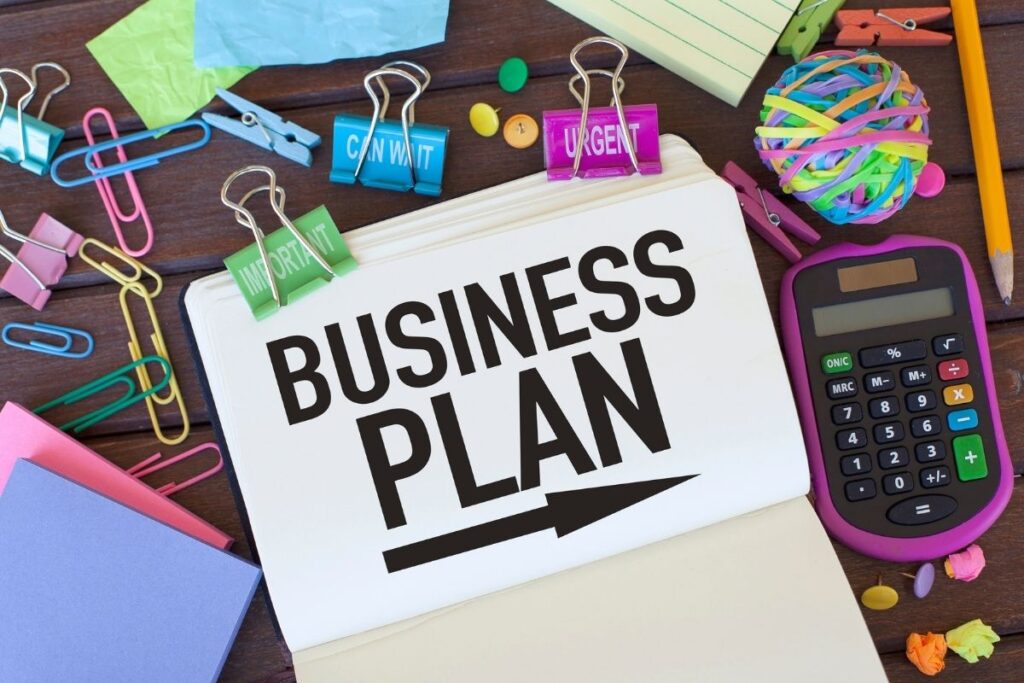 Open notebook with the words “Business Plan” surrounded by colorful binder clips, calculator, and office supplies.