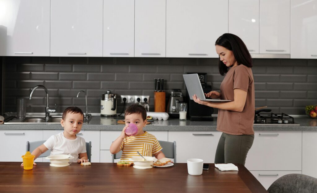 Stay-at-home mom standing in a kitchen using a laptop while two young children eat breakfast at the table.