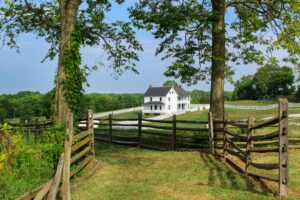 White farmhouse behind a rustic wooden fence on a large green property.