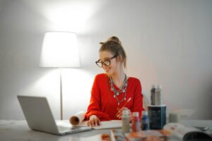 Woman in glasses working from home on a laptop, smiling while taking notes at her desk with office supplies and a lamp.