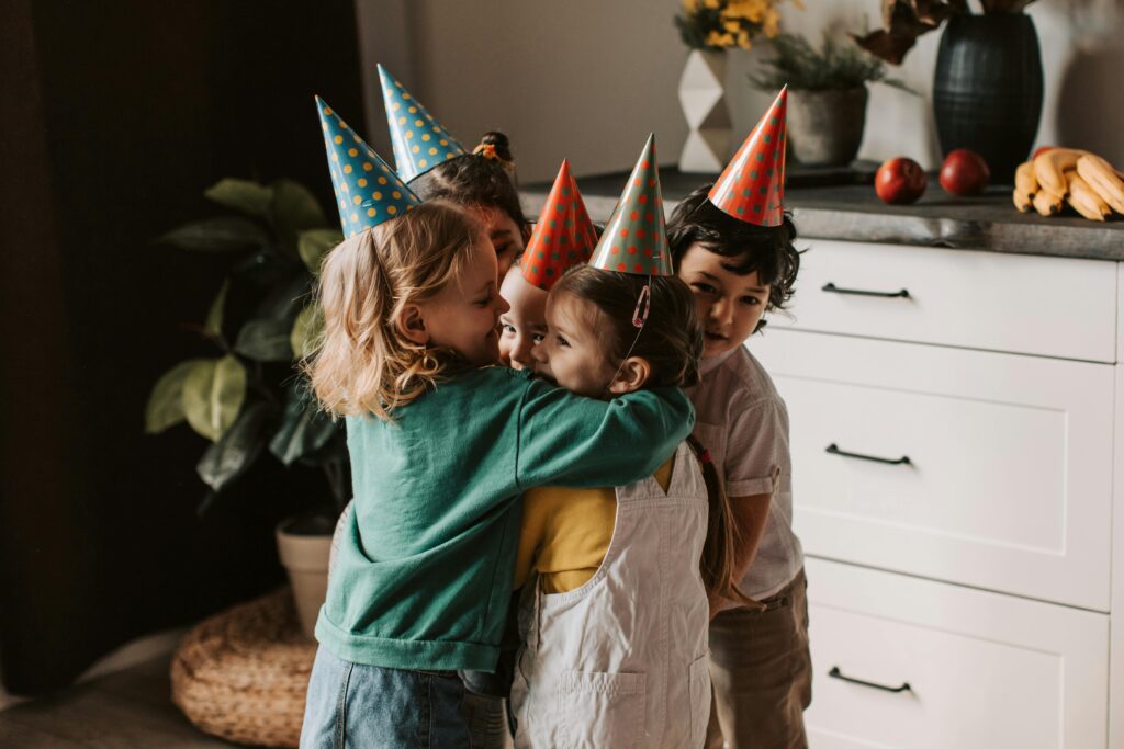 Four children wearing colorful party hats hugging and smiling at a birthday celebration indoors.