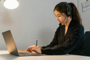 Woman working as a virtual assistant, wearing a headset, typing on a laptop while taking notes at her desk.