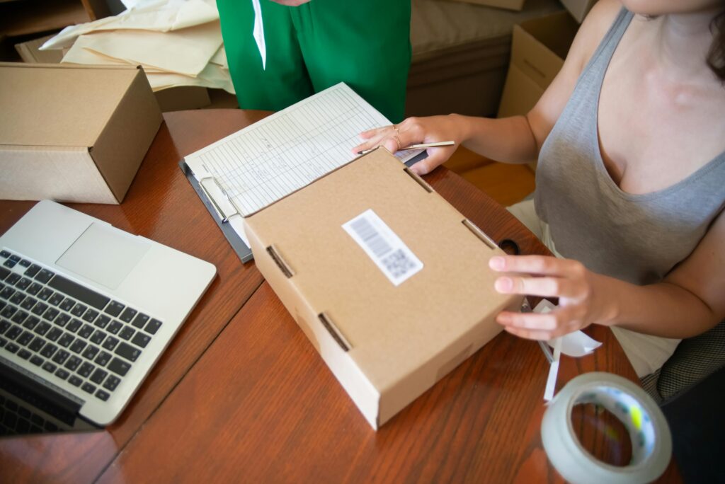 Person preparing an e-commerce order at home, packaging a cardboard shipping box with a laptop and order sheet on the table.