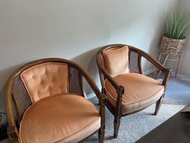 Pair of vintage wooden armchairs with peach cushions and cane backs placed against a neutral wall beside a wicker plant stand.