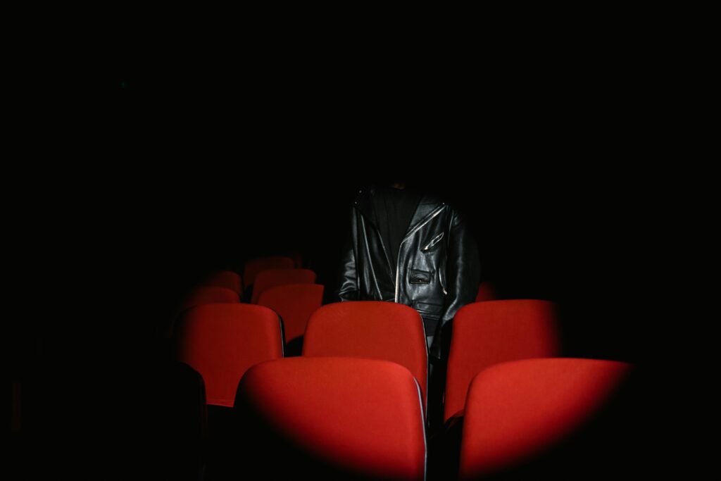 A spotlight shines on empty red theater seats with a figure in a black leather jacket standing in the shadows, symbolizing drama and mystery.