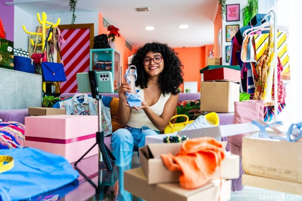 Smiling reseller recording a video while showcasing shoes surrounded by clothing, boxes, and shipping supplies in a colorful home workspace.