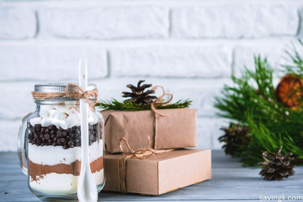 Homemade hot cocoa mix in a mason jar layered with cocoa powder, sugar, chocolate chips, and marshmallows, tied with twine and placed beside rustic wrapped gifts and pine decorations.