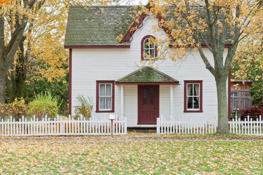 White farmhouse with red trim and picket fence surrounded by autumn trees.