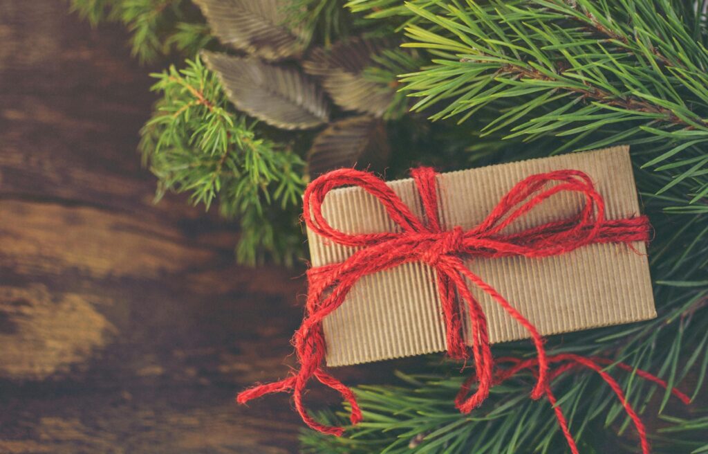Simple brown paper Christmas gift with a red string bow surrounded by pine branches on a wooden table.