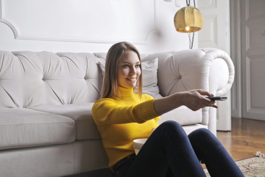 Woman sitting on the floor watching TV at home with a remote in hand and a bowl of snacks.