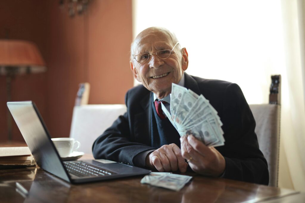 Elderly man in a suit smiling while holding a fan of dollar bills at his desk with a laptop and coffee cup, symbolizing timeless money habits and financial wisdom.