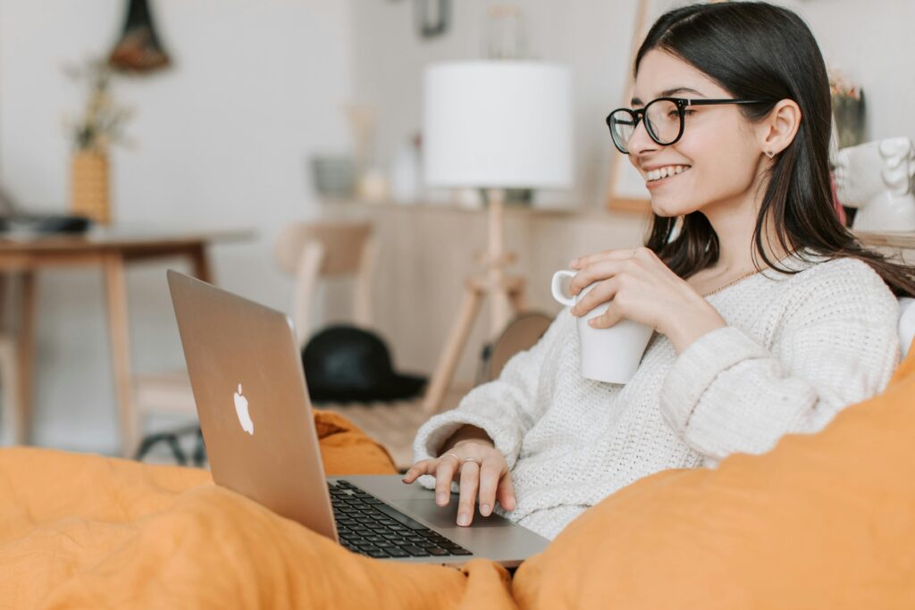 Smiling woman sitting comfortably with a laptop and coffee mug, shopping online from home in a cozy setting.