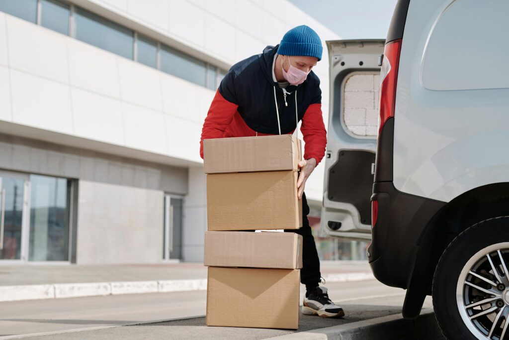 Man wearing a beanie and mask unloading cardboard boxes from a delivery van outside a building.
