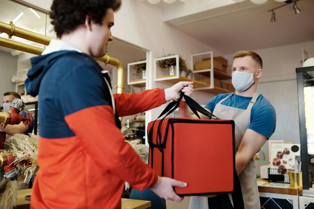 Delivery driver picking up a food order from a masked restaurant worker using an insulated red delivery bag.