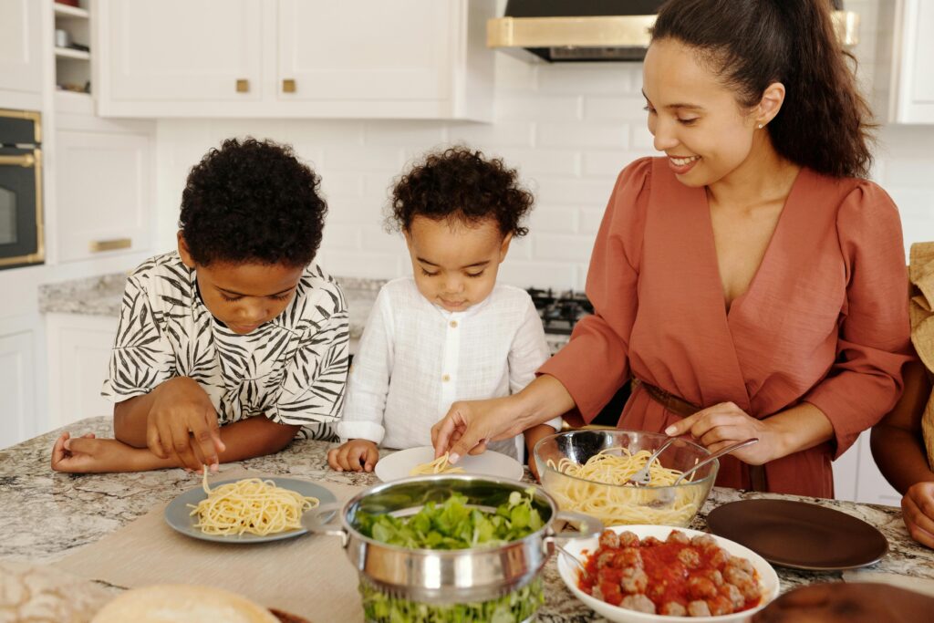 A mother serves spaghetti to her two young children at the kitchen counter, showing how to make simple, affordable meals stretch during tough times.