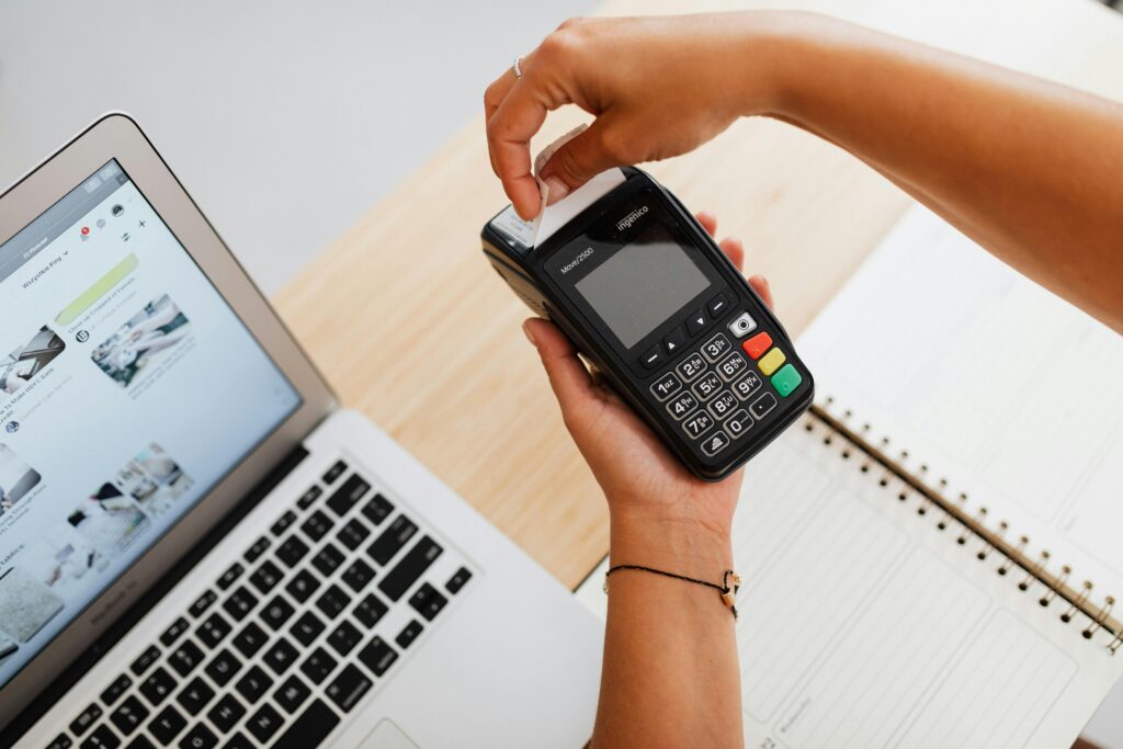 Person processing a card payment using a POS terminal next to a laptop and notebook on a wooden desk.