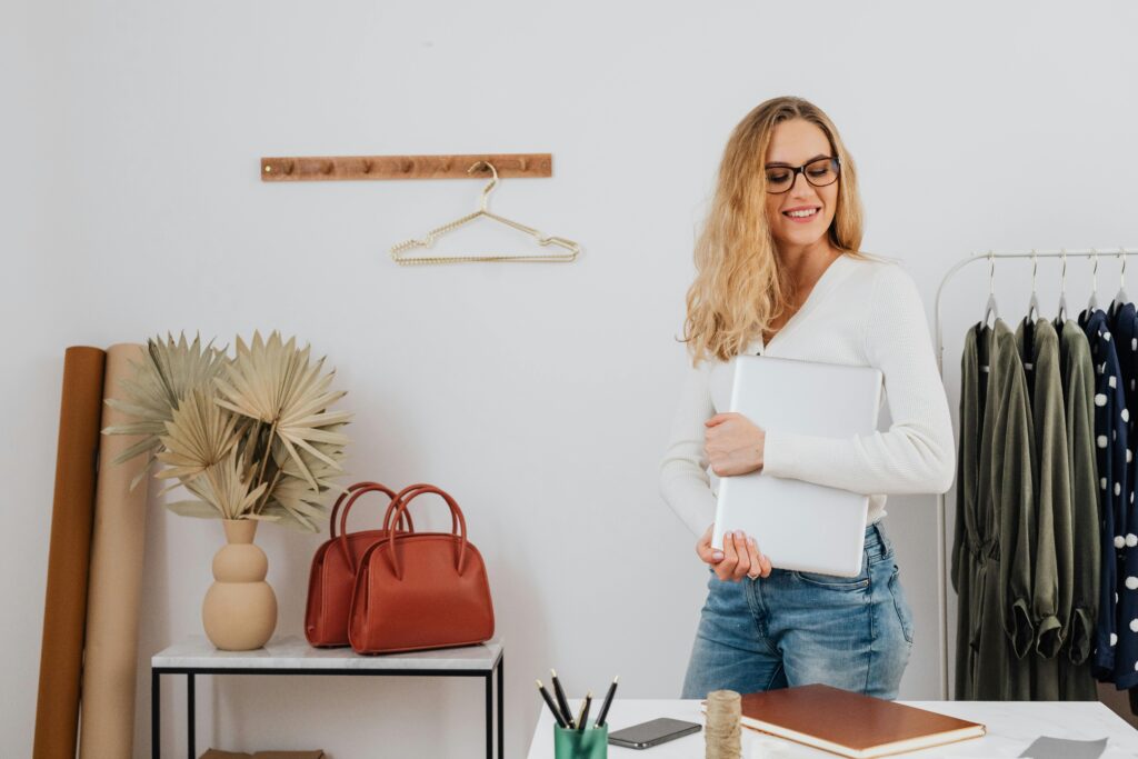 Boutique owner smiling while holding a laptop in a stylish workspace with handbags and clothing racks in the background.