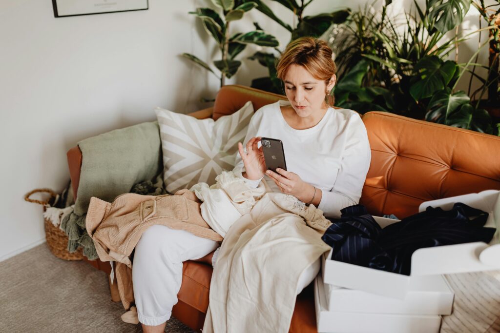 Woman sitting on a couch using her smartphone to find free clothes online, surrounded by clothing and shipping boxes.