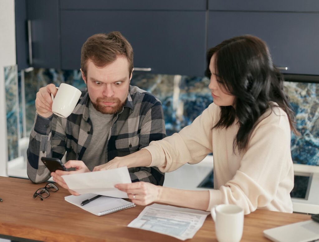 A worried couple reviews bills together at the kitchen table, using a phone and calculator to manage their budget during tough financial times.