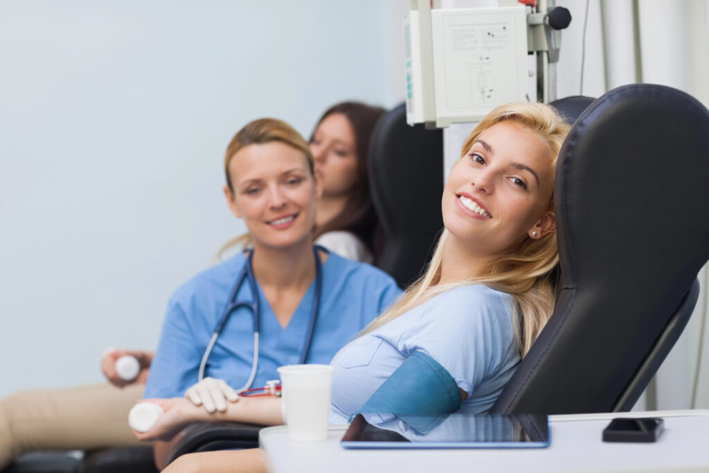 Smiling woman donating plasma while a nurse assists at a medical clinic.