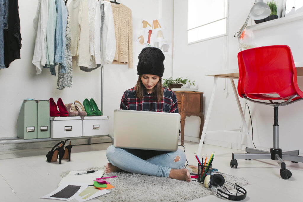 Young blogger working from home on laptop surrounded by clothes, shoes, and creative supplies.