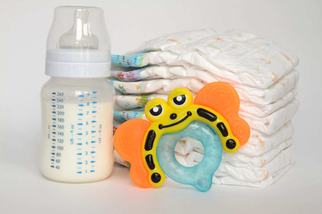 Baby bottle with milk, colorful teething toy, and stack of diapers on a white background.