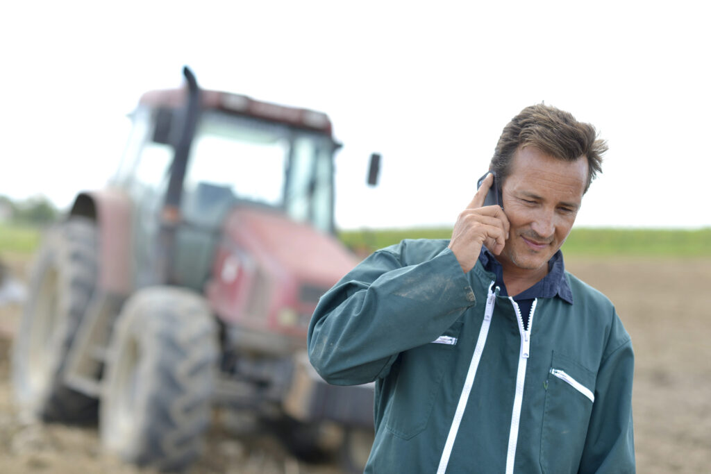 Farmer talking on a cellphone in a field with a red tractor in the background, representing modern side hustles and communication in farming.