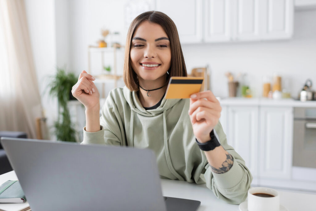 Young woman smiling while holding a credit card and shopping online from her laptop at home.