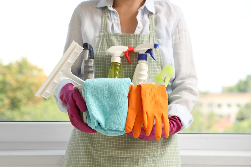 Person wearing apron and gloves holding a basket of cleaning supplies including spray bottles, cloths, and rubber gloves.