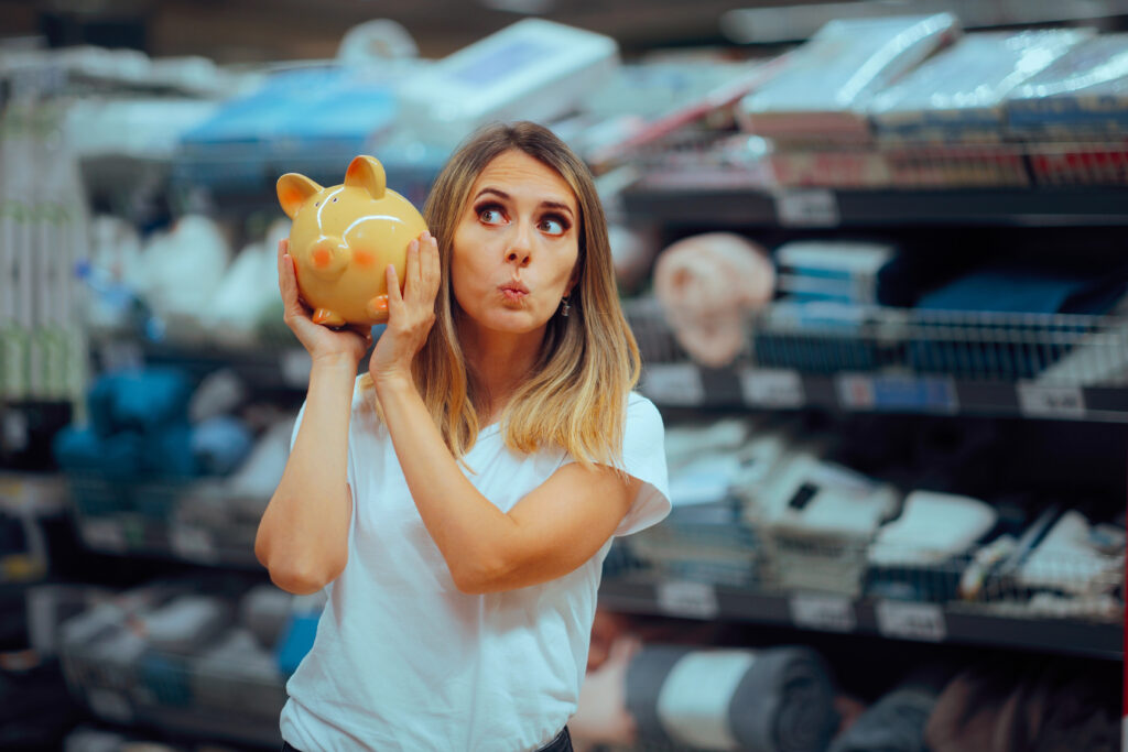 Woman holding a piggy bank to her ear while shopping, symbolizing frugal habits.