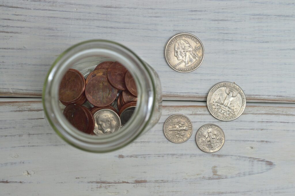 Glass jar filled with coins sitting on a white wooden surface with loose coins scattered around it.