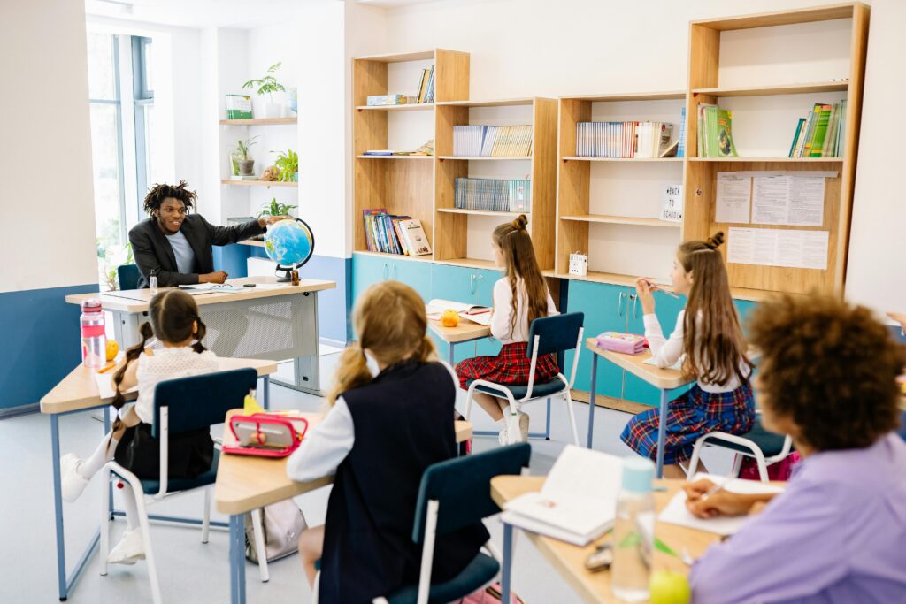 A teacher pointing to a globe while students in school uniforms sit at desks listening attentively in a bright classroom.