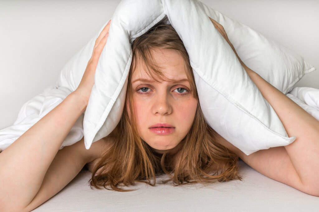 Tired woman lying in bed with a pillow over her head, looking stressed and unable to sleep