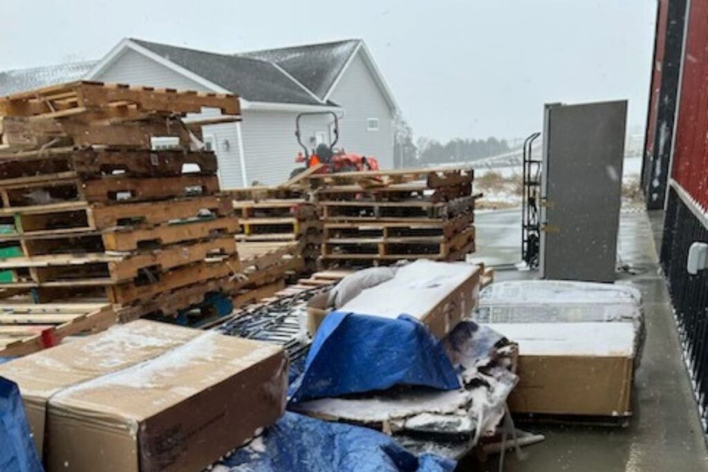 Stacks of free wooden pallets covered lightly in snow behind a warehouse, with boxes, tarps, and outdoor equipment, showing the kind of free pallets people often pick up for projects or resale.