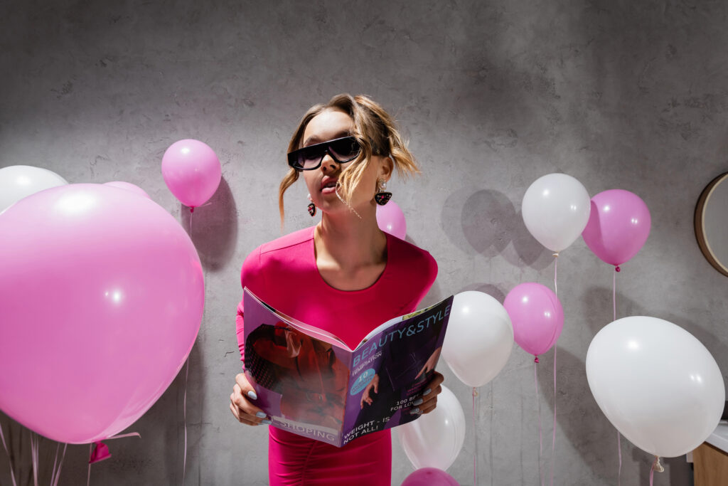 Woman in a pink dress reading a beauty catalog surrounded by pink and white balloons, representing free beauty catalogs by mail