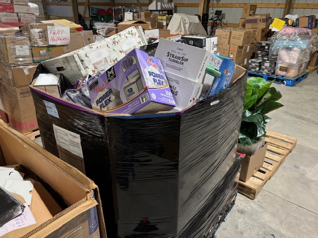 Large wrapped liquidation pallet filled with assorted retail boxes and merchandise inside a warehouse setting.