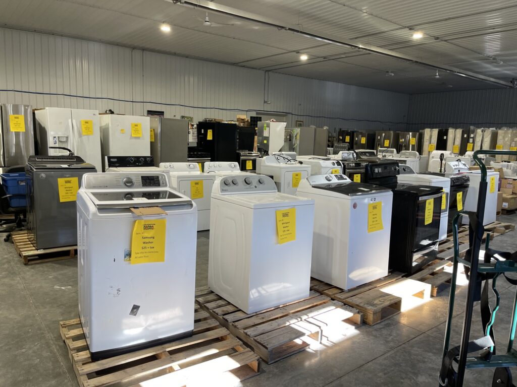 Rows of used washers, dryers, and appliances for sale on pallets inside a liquidation warehouse