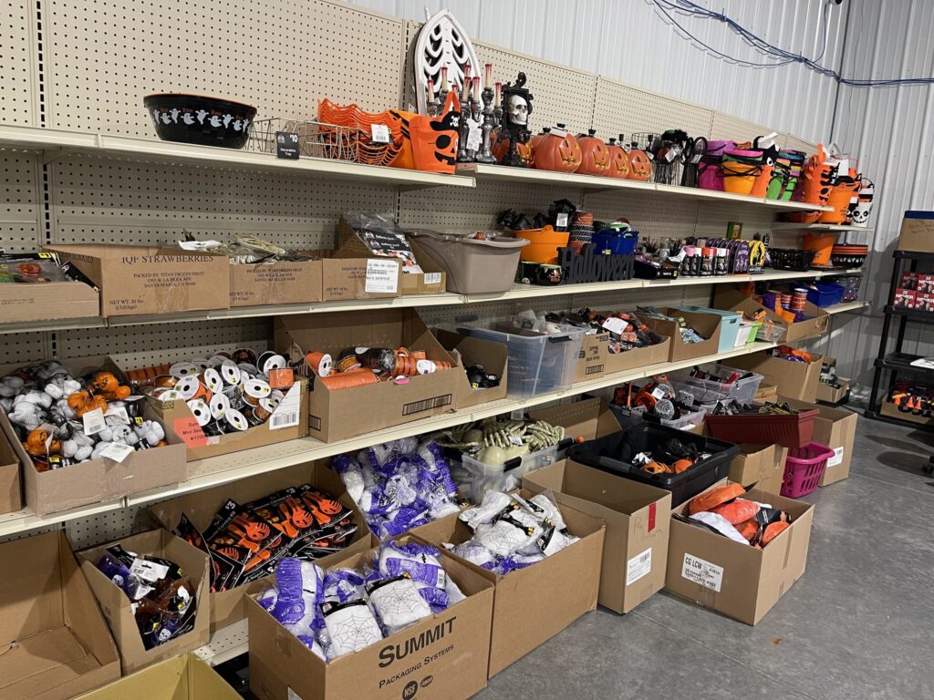 Halloween liquidation shelf with clearance decorations, bins of supplies, and bulk boxes of seasonal items for resale