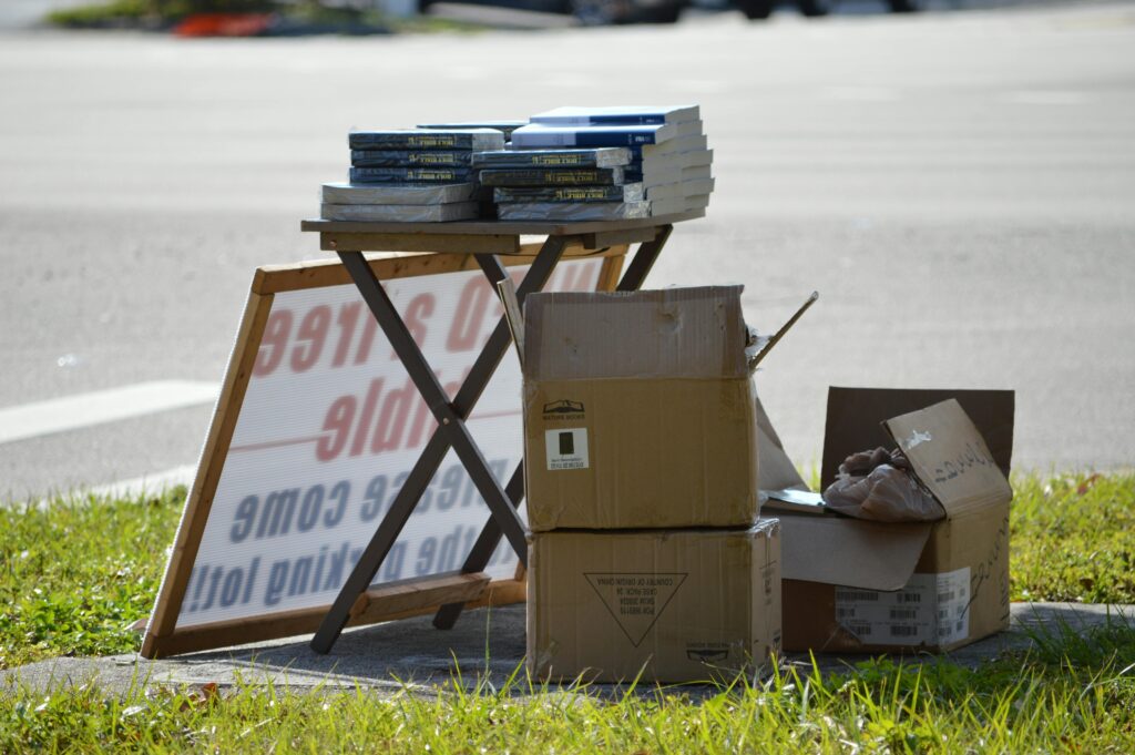 Yard sale items displayed on table with boxes and books for sale outdoors