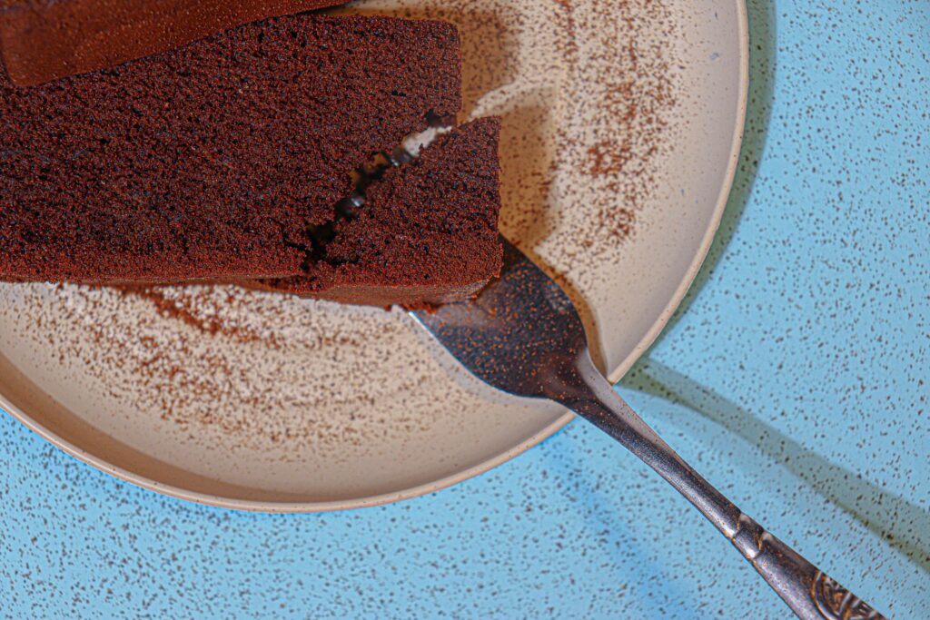 Close-up of a slice of chocolate cake on a plate with cocoa powder dusted on top and a fork beside it on a blue surface.