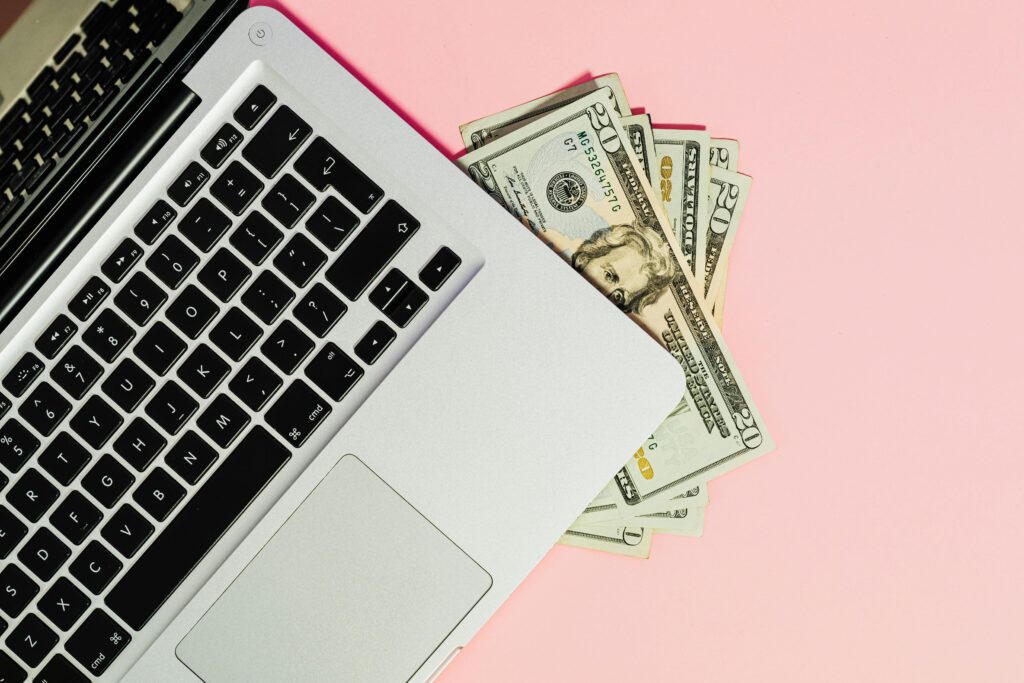 Laptop with US dollar bills tucked underneath on a pink background, representing making money online