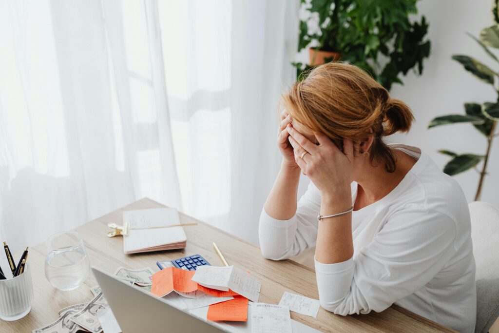 Woman sitting at a table with bills, calculator, and cash, holding her head in stress over finances