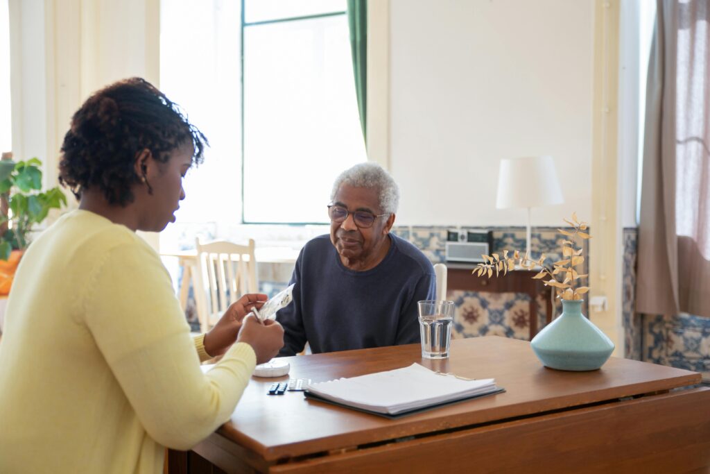 Caregiver helping an elderly man organize medication and healthcare paperwork at a kitchen table at home.