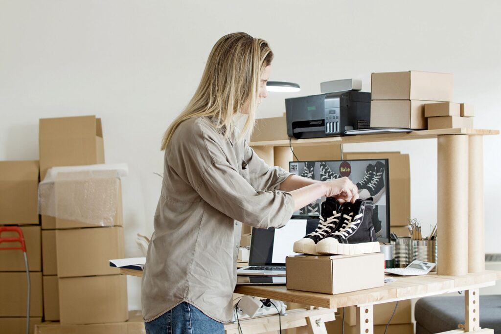 Online reseller photographing a pair of sneakers for sale while packing orders in a home business workspace with shipping boxes and inventory.